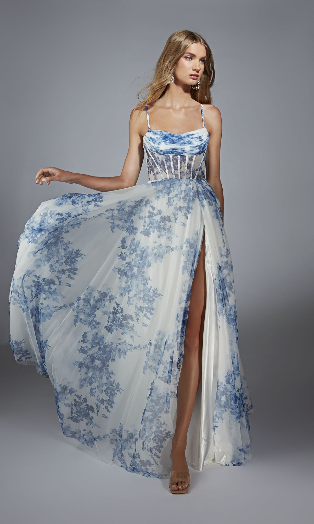 A woman poses against a plain gray background wearing the Alyce 61752 long prom dress, featuring blue floral prints, a corset bodice, and a thigh-high slit.